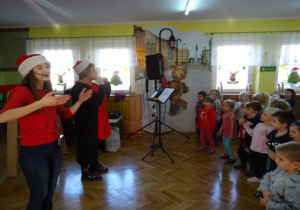 Prowadzące koncert w mikołajowych czapkach i tańczące dzieci.
